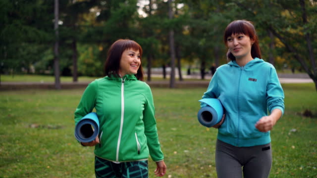 Smiling-young-women-are-walking-in-park-with-yoga-mats-and-talking-sharing-news-and-discussing-practice-together.-Youth,-outdoor-activity-and-conversation-concept.