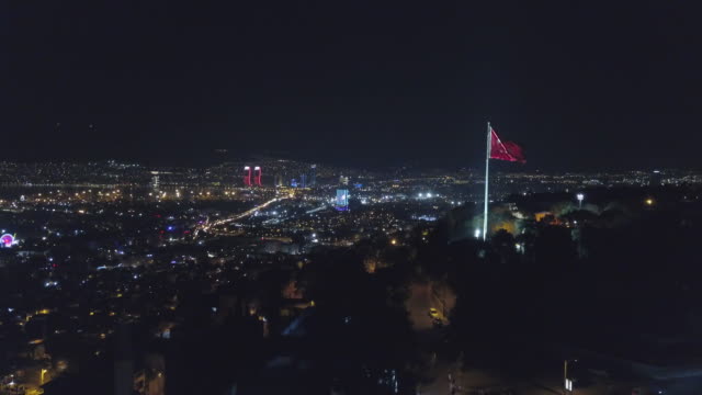 Skyline-der-Stadt-gezeichnet-durch-die-Nacht-Drohne,-Skyline-Stadtflagge-vor-Kadifekale,-izmir