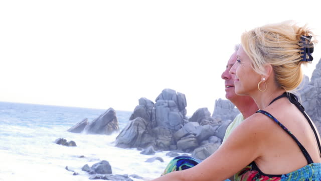 Eine-ältere-paar-sitzen-am-Strand-und-Blick-auf-die-Wellen-und-dann-lächelt-in-die-Kamera