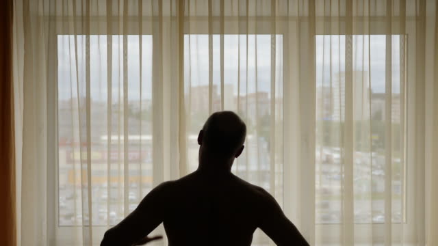 Silhouette-of-a-man-performing-exercise-indoors.-In-the-background-city-street-behind-window-curtains.-Men-warming-up-do-morning-stretching.