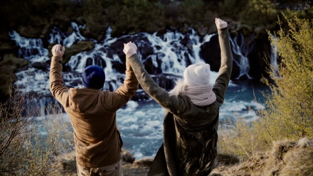 Rückansicht-des-jungen-stilvolles-Paar-Hände-hob-und-fühle-mich-glücklich,-Freiheit-in-der-Nähe-der-Wasserfälle-in-Island