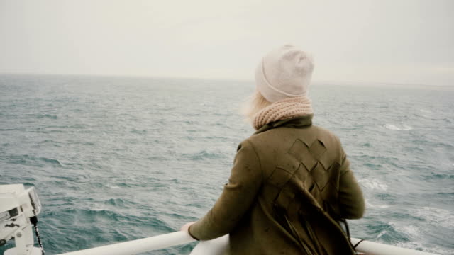 Vista-posterior-de-la-mujer-joven-rubia-de-pie-en-el-barco-y-mirando-con-nostalgia-sobre-el-mar,-explorando-la-Islandia