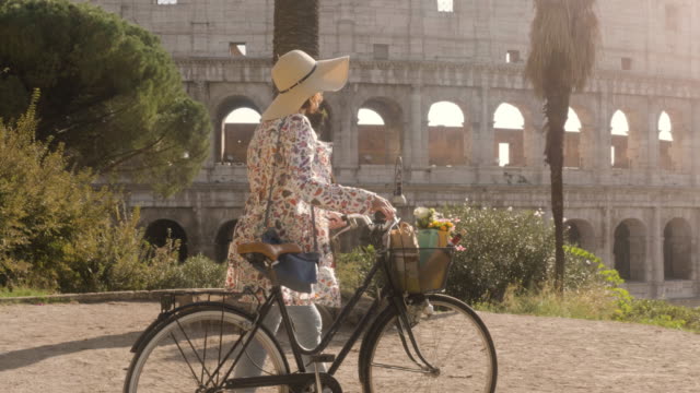 Beautiful-young-woman-in-colorful-fashion-dress-walking-alone-on-hill-with-bike-in-front-of-colosseum-in-Rome-at-sunset-with-trees-attractive-girl-with-straw-hat