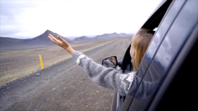 Cheerful-caucasian-female-inside-car-looking-at-road-arms-outstretched-,-mountain-volcanic-landscape.-Road-trip-concept.--Blond-hair-girl-looking-at-landscape.-SLOW-MOTION