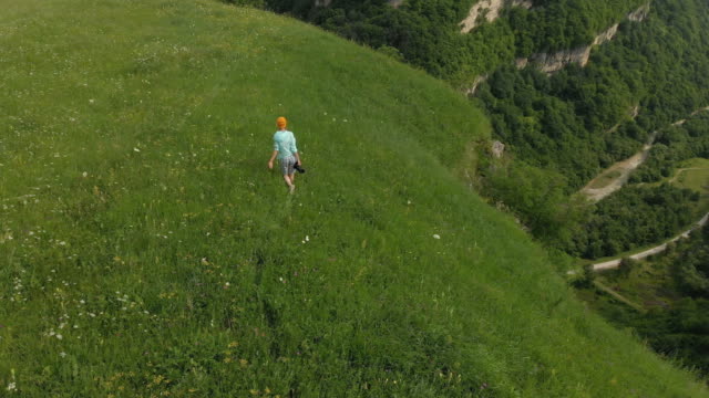 The-girl-photographer-in-glasses-and-a-hat-walks-with-her-dslr-camera-on-the-edge-of-the-plateau-near-the-precipice.-Aerial-view