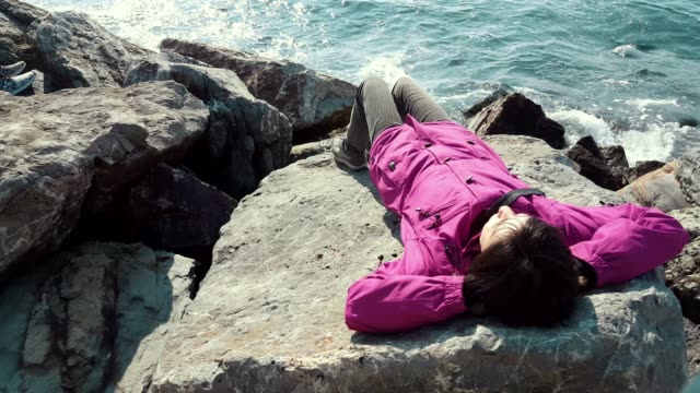 Young-Caucasian-woman-with-pink-coat-lying-on-the-rocks-near-the-wavy-sea