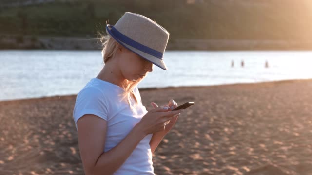 Schönes-Blondes-Mädchen-mit-Hut-Typen-eine-Nachricht-auf-ihr-Handy-am-Strand-bei-Sonnenuntergang.