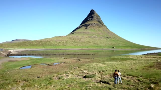 Drone-shot-aerial-view-of-caucasian-couple-contemplating-the-famous-Kirkjufell-mountain-peak,-nature-background-with-grass-and-mountains.-Shot-in-West-Iceland,-Springtime.-People-travel-lifestyles-concept--4K-video