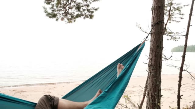 Young-caucasian-male-resting-lying-on-blue-hammock-outdoors-near-lake.