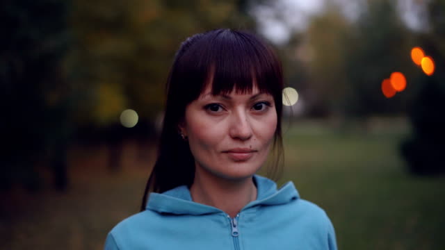 Portrait-of-beautiful-young-sportswoman-with-long-dark-hair-and-brown-eyes-looking-at-camera-and-smiling-standing-outdoors-in-autumn-evening-wearing-hoody.