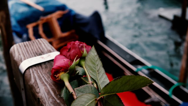 Close-up-shot-of-beautiful-red-roses-lying-on-wooden-canal-pier,-traditional-Italian-gondola-rocking-on-waves-nearby.