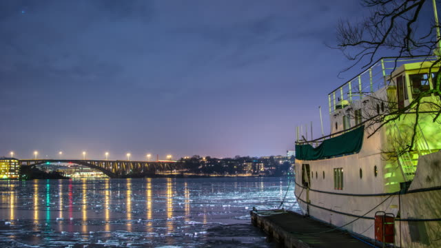 Bridge-and-frozen-bay-at-night-4K-Time-Lapse-Tilt.-Quay-and-boat,-lights-reflected-on-the-ice