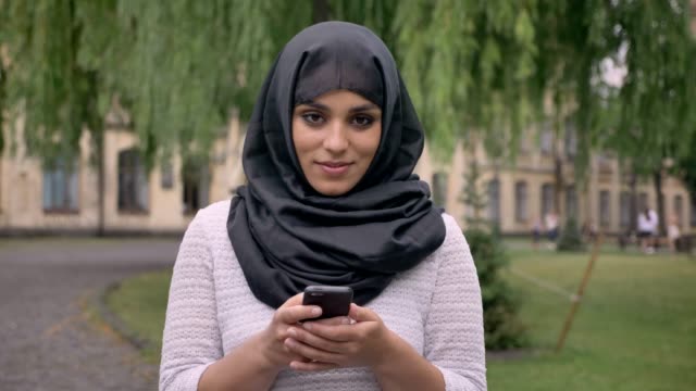 Young-beautiful-muslim-girl-in-hijab-is-typing-message-on-her-smartphone,-watching-at-camera-and-smiling-in-blue-weather,-communication-concept,-religious-concept