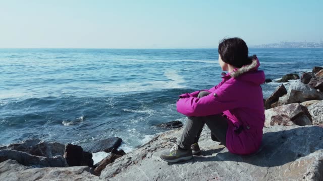 Mujer-joven-Caucásica-con-capa-rosa,-sentada-en-las-rocas-cerca-del-mar-ondulado