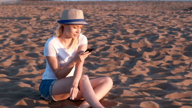 Hermosa-chica-rubia-en-un-sombrero-sentado-en-la-playa-al-atardecer-con-el-teléfono-móvil.