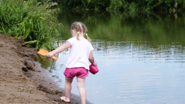 Little-girl-is-walking-with-a-toy-spatula-and-a-bucket-in-the-sand-near-the-river