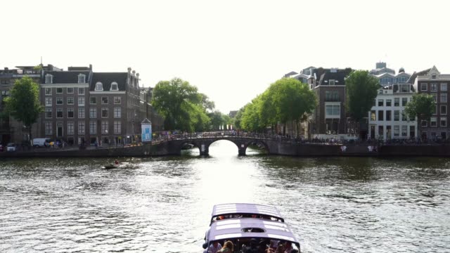 Canal-cruise-tour-boat-sails-in-iconic-canal-with-traditional-bridge-in-Amsterdam,-Holland-Europe