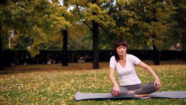 Flexible-young-lady-is-doing-exercises-for-healthy-spine-sitting-on-yoga-mat-and-moving-back-and-body-during-individual-practice-in-park.-Health-and-nature-concept.