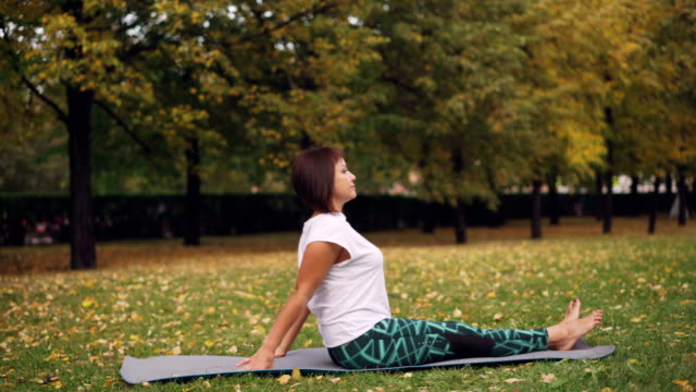 Hermosa-chica-en-ropa-de-deporte-es-ejercicio-en-el-Parque-practicando-giros-sentado-y-adelante-curvas-en-estera-de-yoga-cálido-día-de-otoño.-Concepto-de-naturaleza,-deportes-y-salud.