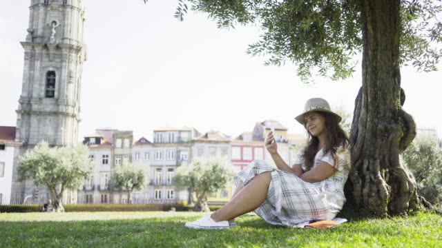 Woman-in-dress-with-smartphone-sitting-under-tree-in-park