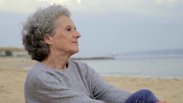 Thoughtful-senior-lady-sitting-on-beach-and-looking-at-distance.