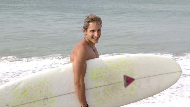 SLOW-MOTION:-Smiling-young-surfer-walking-along-the-beach