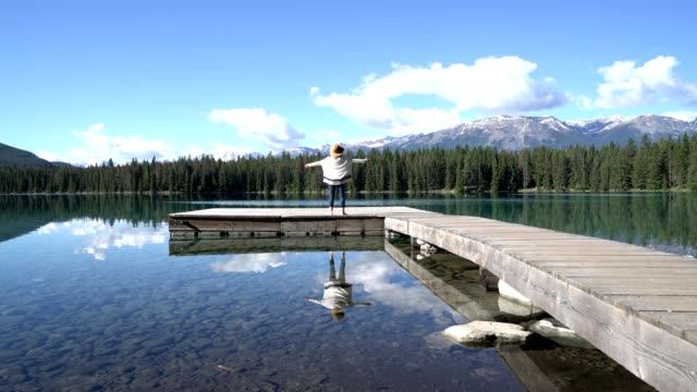 Young-woman-walks-on-wooden-pier-above-stunning-mountain-lake-scenery,-arms-wide-open