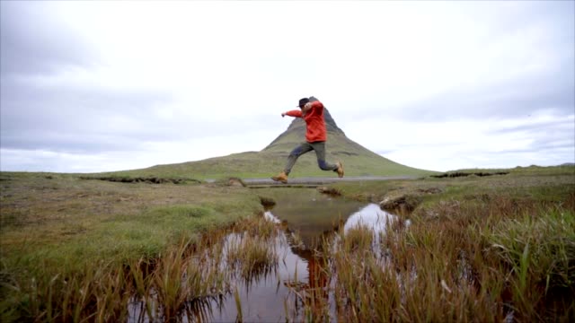 Junger-Mann-in-Island-über-Fjord-Fluss-am-berühmten-Berg-Kirkjufell-springen
