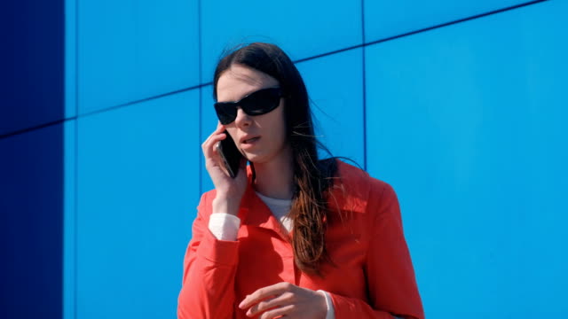 Mujer-morena-joven-en-gafas-de-sol-habla-en-el-teléfono-al-lado-del-edificio-azul.