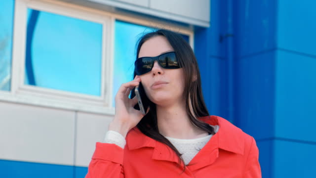 Portrait-von-junge-Brünette-Frau-mit-Sonnenbrille-und-roten-Mantel-spricht-am-Telefon-neben-blauen-Gebäude.