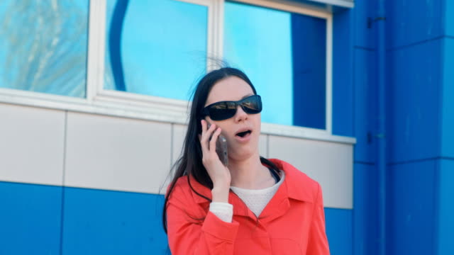 Portrait-of-young-brunette-woman-in-sunglasses-and-red-coat-speaks-on-the-phone-beside-blue-building.
