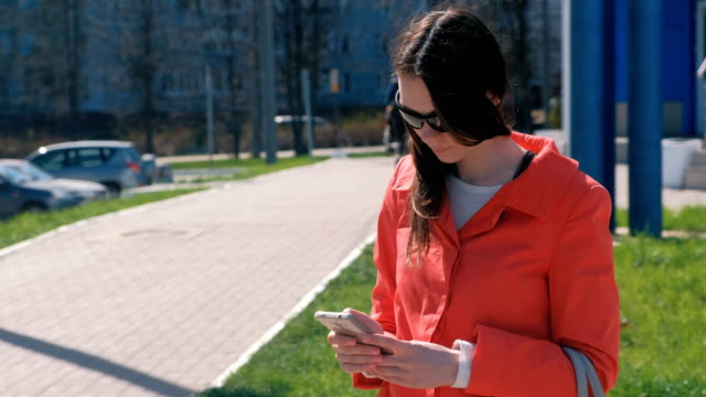Brünette-junge-Frau-mit-Sonnenbrille-im-roten-Mantel-wartet-auf-jemanden-und-überprüft-ihr-Handy-SMS.