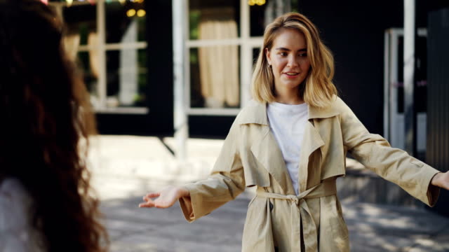 Attractive-girl-is-talking-to-her-friend-standing-in-the-street-in-the-city-and-gesturing-sharing-news.-Emotional-young-woman-with-blond-hair-is-wearing-trendy-coat.