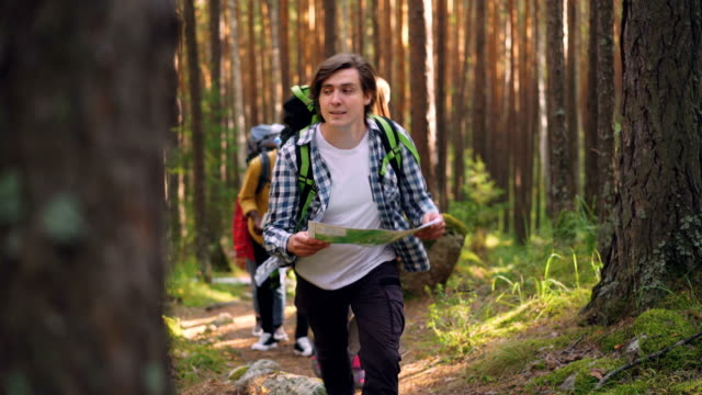 Hübscher-Kerl-hält-Karte-und-gehen-im-Wald,-während-seine-Freunde-ihm-auf-Trail-im-Holz-folgen.-Reisen,-Sommer-und-Orientierung-Konzept.