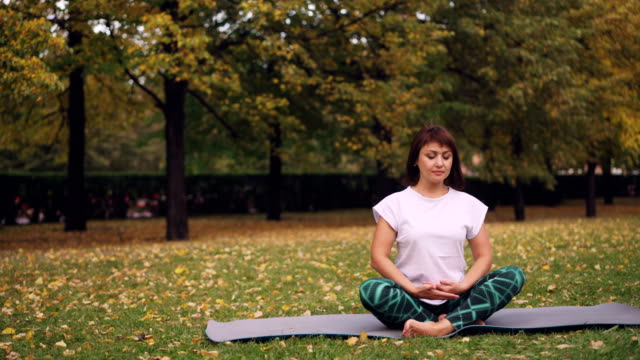Pretty-young-woman-is-relaxing-sitting-in-lotus-pose-on-yoga-mat-in-park-and-breathing-resting-after-individual-practice.-Meditation-and-nature-concept.
