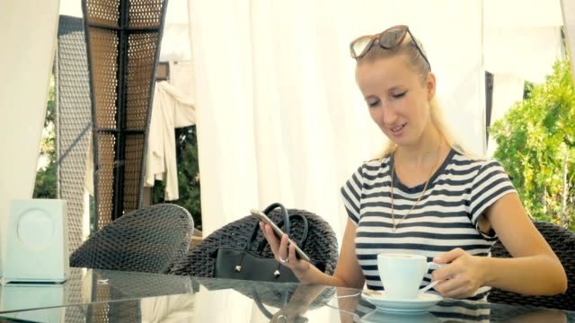 Young-girl-is-sitting-in-a-summer-cafe,-drinking-delicious-coffee-and-writing-a-message-on-the-phone.