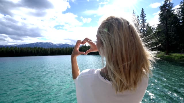 Young-woman-making-heart-shape-finger-frame-on-mountain-lake