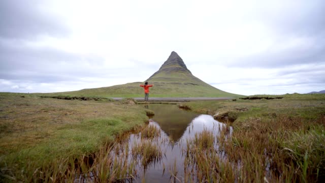 4K---Young-man-in-Iceland-arms-outstretched-for-freedom-Springtime-overcast-sky-at-famous-Kirkjufell-mountain