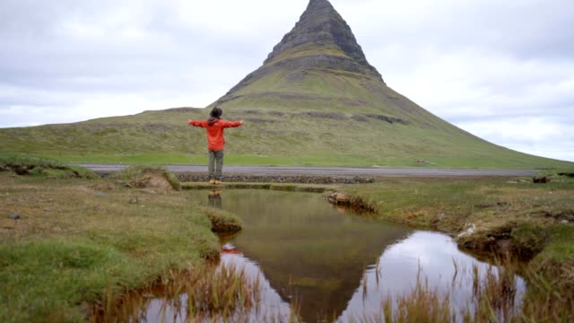 4K---junger-Mann-in-Island-Arme-ausgestreckt-für-Freiheit-Frühling-bedeckten-Himmel-am-berühmten-Berg-Kirkjufell