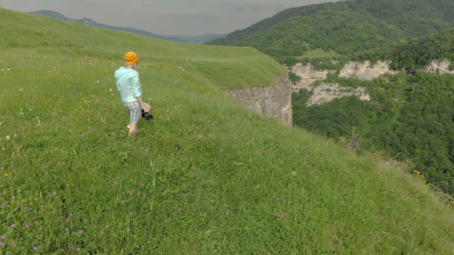 The-girl-photographer-in-glasses-and-a-hat-walks-with-her-dslr-camera-on-the-edge-of-the-plateau-near-the-precipice.-Aerial-view