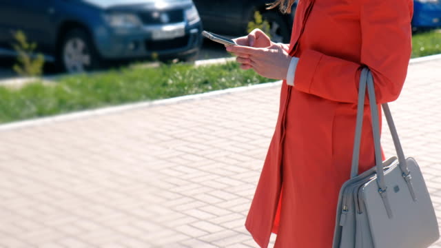 Nicht-erkennbare-Frau-im-roten-Mantel-wartet-auf-jemanden-und-überprüft-ihr-Handy-SMS.-Close-up-Hände.-Seitenansicht.