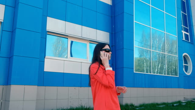 Porträt-der-glückliche-junge-Brünette-Frau-mit-Sonnenbrille-am-Telefon-spricht-die-neben-blauen-Gebäude.