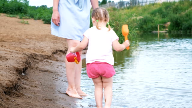Small-child-is-playing-in-the-river,-the-girl-pours-water-out-of-the-pot-in-the-summer.-Mom-looks-after-her-daughter.