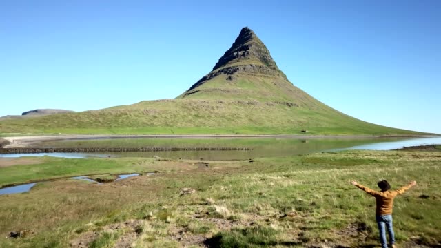 Drone-shot-aerial-view-of-Caucasian-male-arms-raised-for-positive-emotions,-Kirkjufell-mountain-on-background.-Shot-in-West-Iceland,-Springtime.-People-travel-carefree-lifestyles-concept