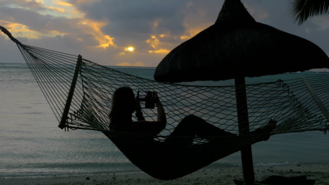 Mujer-relajante-en-hamaca-en-la-playa-y-toma-fotos-de-celular