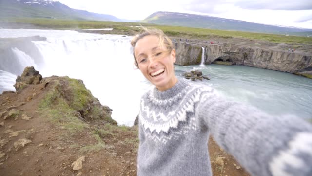 Young-woman-taking-selfie-portrait-with-magnificent-waterfall-in-Iceland,-Godafoss-falls.-People-travel-exploration-concept