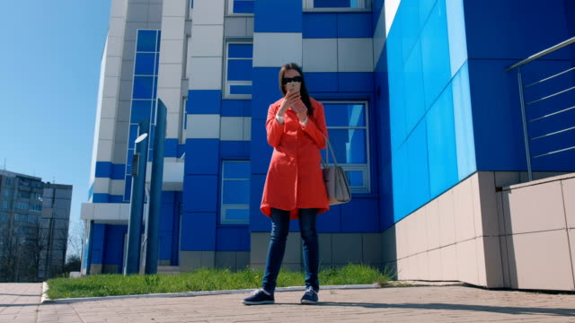 Brünette-junge-Frau-mit-Sonnenbrille-im-roten-Mantel-wartet-auf-jemanden-und-überprüft-ihr-Handy-SMS.