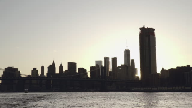 Una-puesta-de-sol-sunflare-brilla-a-través-de-skyline-de-Lower-Manhattan-y-el-puente-de-Brooklyn-en-Nueva-York,-Estados-Unidos-filmado-desde-el-East-River