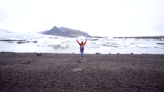 Tourist-travel-man-stands-arms-outstretched-by-glacier-lagoon-in-Iceland