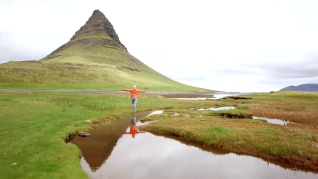Drone-view-aerial-of-Young-woman-in-Iceland-arms-outstretched-for-freedom-Springtime-overcast-sky-at-famous-Kirkjufell-mountain--4K-footage
